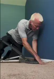 Carpet technician using a power stretcher to re-tension and smooth a plush beige residential carpet.